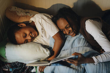 Happy siblings reading story book while lying on bed at home - MASF30285