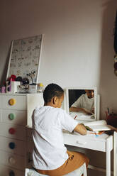 Rückansicht eines Jungen, der in ein Buch schreibt, während er zu Hause am Tisch im Arbeitszimmer sitzt - MASF30287