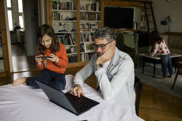 Freiberufler, der an einem Laptop arbeitet, während seine Tochter ein Smartphone auf dem Tisch benutzt und ein Mädchen im Hintergrund spielt - MASF30322