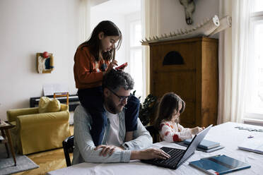 Daughter sitting on shoulder of father working on laptop at home - MASF30326