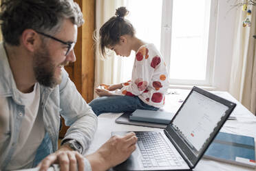 Lächelnder Mann arbeitet am Laptop, während seine Tochter am Tisch ihr Smartphone benutzt - MASF30328