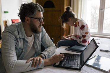 Freiberufler, der an einem Laptop arbeitet, während seine Tochter ein Mobiltelefon benutzt und zu Hause auf dem Tisch sitzt - MASF30329