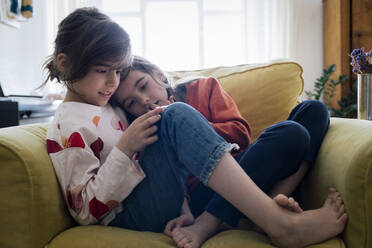 Mädchen mit Smartphone sitzt mit Schwester auf Sessel im Wohnzimmer zu Hause - MASF30332