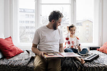 Happy man looking at daughter sitting with piano on bed at home - MASF30334