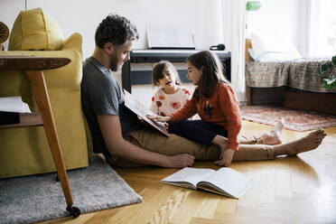Girl looking at sister reading book sitting with father in living room at home - MASF30335