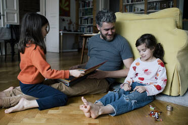 Smiling man looking at daughter playing with toys while girl reading book at home - MASF30336