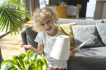 Happy girl holding watering can by sofa at home - SVKF00188