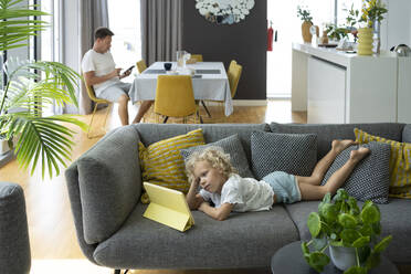 Mädchen schaut Tablet-PC auf Sofa mit Vater auf Stuhl im Wohnzimmer zu Hause sitzen - SVKF00200
