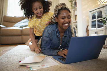 Glückliche Tochter beobachtet Mutter bei der Arbeit am Laptop auf dem Wohnzimmerboden - CAIF32722