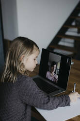 Girl writing on paper studying through video call on laptop at home - LHPF01454