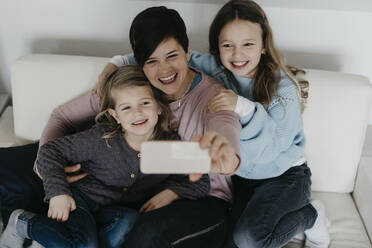 Smiling mother taking selfie with daughters sitting on sofa at home - LHPF01458