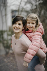 Lächelnde Frau mit kurzen Haaren, die ihre Tochter im Hinterhof huckepack nimmt - LHPF01461