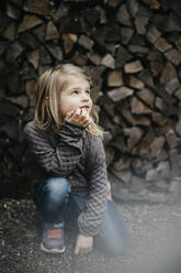 Thoughtful girl with hand on chin kneeling in front of stack of wood - LHPF01462