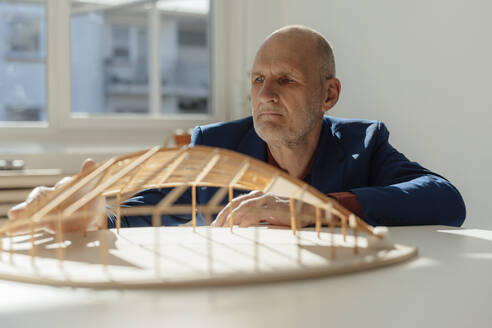 Senior architect analyzing leaf shape wooden model at desk in office - JOSEF09685