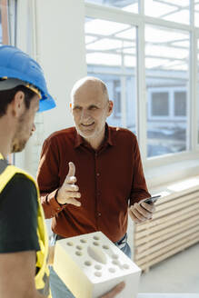 Lächelnder Architekt mit Handy in der Hand, der mit einem Kollegen über einen Betonstein im Büro diskutiert - JOSEF09737