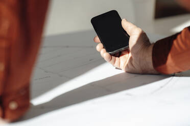 Hand eines Geschäftsmannes, der ein Smartphone auf einer Blaupause im Büro hält - JOSEF09788