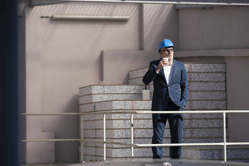 Architekt mit Schutzhelm, der einen Einwegbecher hält, mit der Hand in der Tasche auf einer Baustelle - JOSEF09830