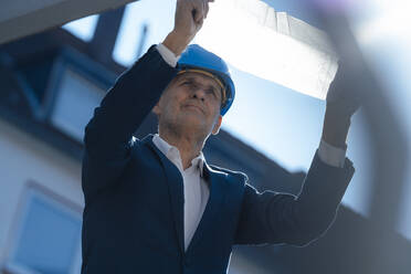 Senior businessman wearing hardhat analyzing blueprint at construction site - JOSEF09839