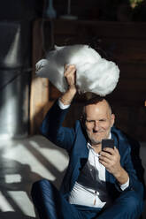 Lächelnder Geschäftsmann, der ein Mobiltelefon hält und eine Wolke über dem Kopf trägt, im Büro - JOSEF09875