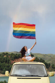 Lächelnde junge Frau mit Regenbogenfahne auf einem Lieferwagen sitzend - DAMF00933