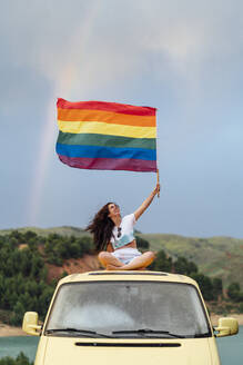 Smiling young woman holding rainbow flag sitting on van - DAMF00933