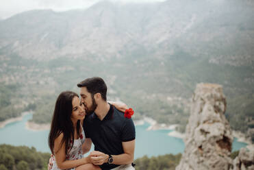 Glückliche Frau, die eine Blume trägt, während ein Mann die Wange küsst und auf dem Gipfel eines felsigen Berges mit malerischer Aussicht sitzt - ADSF34824