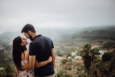 Back view of couple hugging and looking at each other while standing at foggy countryside on summer day - ADSF34832