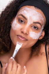 Self assured young ethnic female model with curly dark hair applying mask on face with brush and looking at camera against white background - ADSF34836
