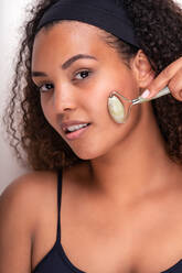 Content young ethnic female with perfect skin and long dark curly hair and headband looking at camera while doing face massage with jade rollers against white background - ADSF34837