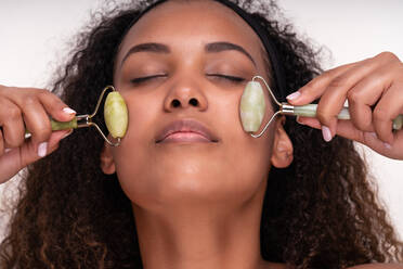 Content young ethnic female with perfect skin and long dark curly hair closing eyes while doing face massage with jade rollers against white background - ADSF34838
