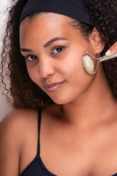 Content young ethnic female with perfect skin and long dark curly hair and headband looking at camera while doing face massage with jade rollers against white background - ADSF34839