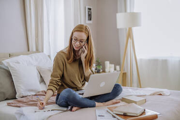 Smiling woman talking on mobile phone writing in book sitting with laptop on bed at home - HAPF03114