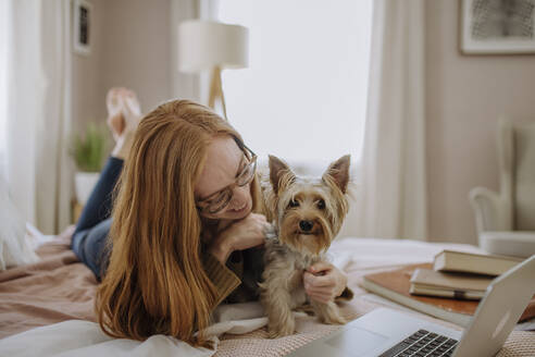 Lächelnde Frau spielt mit ihrem Hund, der zu Hause auf dem Bett liegt - HAPF03133