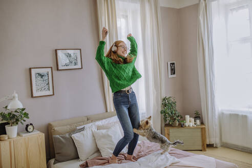 Glückliche Frau hört Musik über drahtlose Kopfhörer und tanzt mit erhobenen Armen auf dem Bett zu Hause - HAPF03134