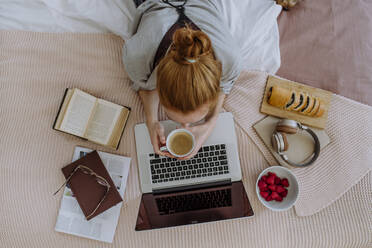 Frau mit Kaffeetasse auf dem Bett liegend und zu Hause am Laptop studierend - HAPF03147