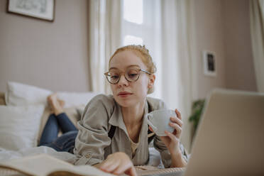 Frau hält Kaffeetasse und liest ein Buch auf dem Bett liegend zu Hause - HAPF03150