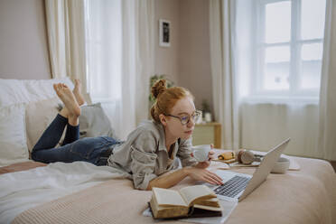 Woman wearing eyeglasses holding coffee cup using laptop lying on bed at home - HAPF03152