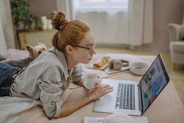 Woman with coffee cup using laptop lying on bed at home - HAPF03156