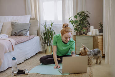 Lächelnde Frau, die sich ein Online-Tutorial über einen Laptop ansieht, während sie auf einer Übungsmatte sitzt und ihren Hund zu Hause hält - HAPF03165