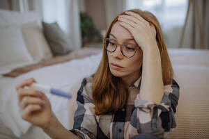 Besorgte Frau mit Brille sitzt mit einem Schwangerschaftstest-Kit vor dem Bett zu Hause - HAPF03180