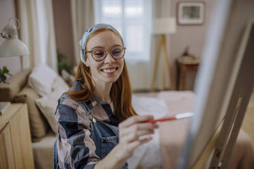 Happy woman wearing eyeglasses and bandana practicing painting at home - HAPF03187