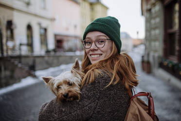 Happy redhead woman wearing knit hat carrying Yorkshire Terrier in city - HAPF03192