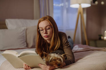 Redhead woman wearing eyeglasses reading book lying with dog on bed at home - HAPF03204