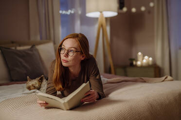 Nachdenkliche Frau mit Brille liegt mit einem Buch neben ihrem Hund auf dem Bett zu Hause - HAPF03206