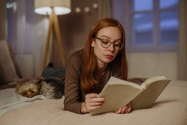 Frau mit Brille liest ein Buch und liegt neben einem Yorkshire Terrier auf dem Bett zu Hause - HAPF03207