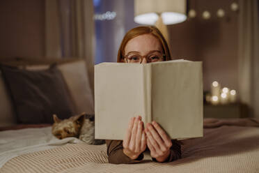 Woman wearing eyeglasses covering face with book on bed at home - HAPF03208