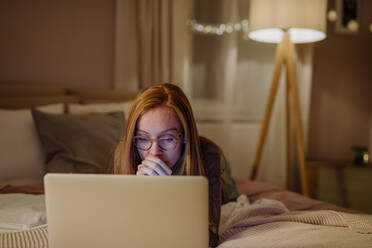 Woman wearing eyeglasses using laptop lying on bed at home - HAPF03217
