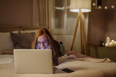 Happy woman with laptop lying on bed at home - HAPF03218