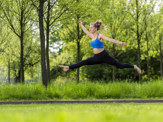 Smiling woman jumping with arms outstretched over meadow at park - STSF03226