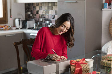 Smiling young woman writing on gift tag for Christmas present at home - EIF04110